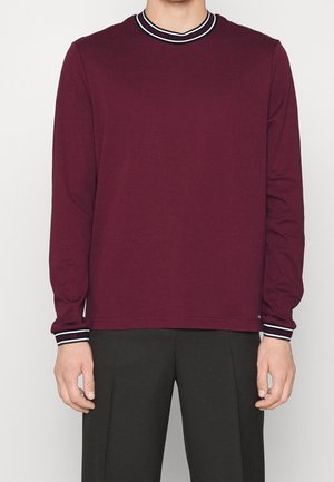 Homme portant une chemise bordeaux à manches longues avec col et poignets rayés noirs et blancs, associée à un pantalon noir.