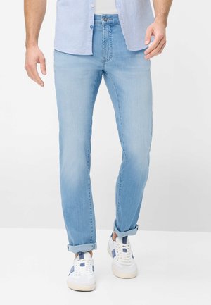 Mand iført lyseblå opkørte jeans, hvide sneakers med blå accenter, lyseblå stribet skjorte og hvid undertrøje, stående på hvidt gulv.