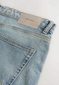 Šviesiai mėlyni džinsai su tiesiu kirpimu, turintys pilką odinį etiketę su užrašu "DENIM SUPPLY" ir matomomis siuvimo detalėmis.