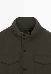 Giacca verde oliva scuro con colletto alto, chiusura a bottoni a pressione e due tasche sul petto. Tessuto liscio, design semplice e senza motivi visibili.