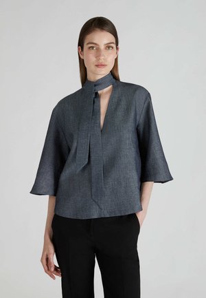 Dunkelgraue Bluse mit einer Schleifenöffnung und halben Ärmeln. Sie hat eine V-förmige Öffnung vorne und ein strukturiertes Gewebe. Kombiniert mit schwarzen Hosen.