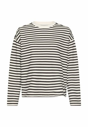 Kvinde står iført sorte jeans med høj talje, sort-hvidstribet langærmet top og hvide sneakers mod en ensfarvet baggrund.
