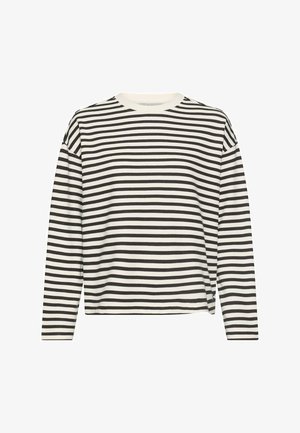 Frau steht, trägt schwarze High-Waist-Jeans, schwarz-weiß gestreiftes langärmliges Oberteil und weiße Sneakers vor einem einfarbigen Hintergrund.