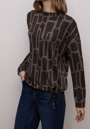Femme portant un haut à manches longues marron foncé avec des motifs géométriques abstraits et un jean bleu, une main dans la poche, sur un fond uni.