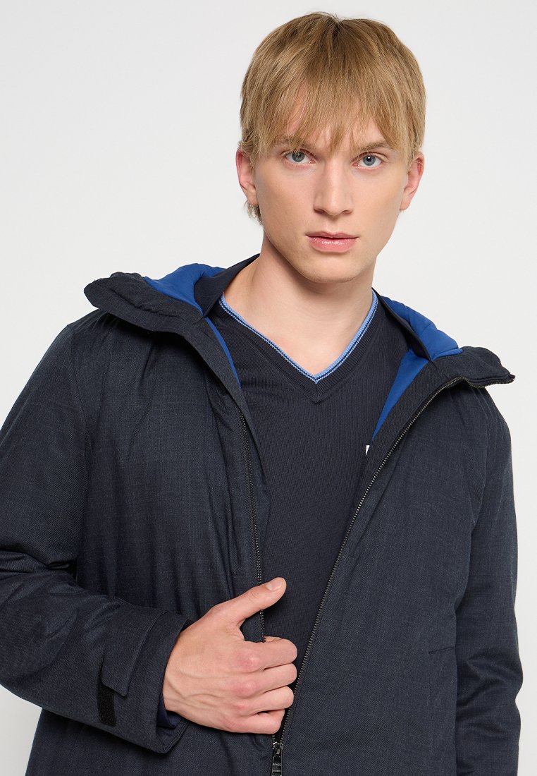 Veste bleu foncé avec capuche, en tissu texturé. En dessous, un t-shirt noir à col en V avec une accentuation bleue au niveau de l'encolure.