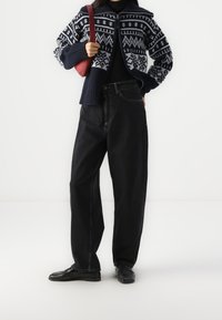 Námořnický pletený kardigan s bílými geometrickými vzory, v kombinaci s černými džíny s vysokým pasem a černými botami. Součástí je červená kabelka.