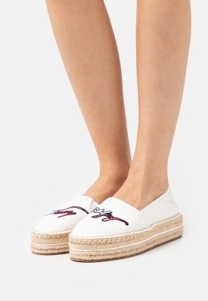 Pieds portant des espadrilles à plateforme blanches avec un motif brodé rouge et bleu sur les orteils, sur un fond blanc uni.