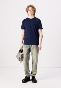 Camiseta de algodón azul marino con mangas cortas, combinada con unos vaqueros verde claro. El modelo sostiene un bolso gris y lleva puestos unos zapatos negros de suela gruesa.
