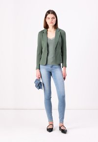 Zelený blazer přes šedý top s krajkovým lemem, v kombinaci s světle modrými skinny džíny a černými balerínami. Modelka drží modrý doplněk.