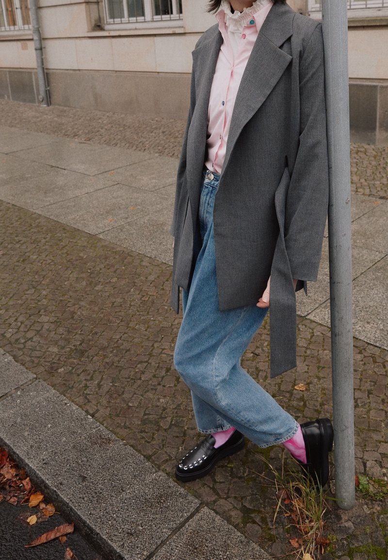 Gray oversized blazer over a pink ruffled shirt, paired with light blue jeans and black shoes, featuring silver studs. Textured pavement background.
