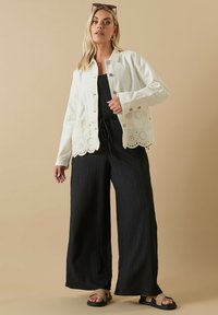 Vitt bomulljacka med öglestickning vid fållen, parat med lösa svarta plisserade byxor och beige sandaler. Modellen står mot en neutral bakgrund.