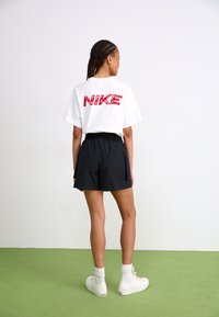 White t-shirt featuring red "NIKE" logo on the back, paired with black shorts and white high-top sneakers. Short sleeves and relaxed fit.