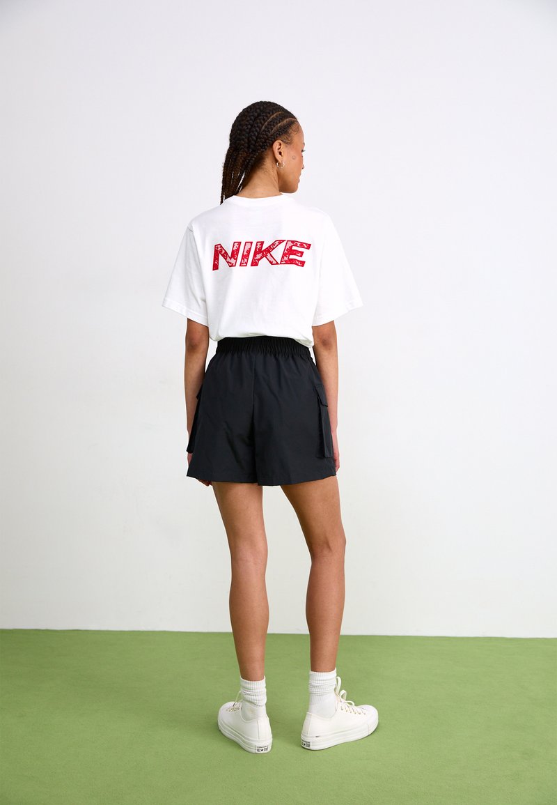 Weißes T-Shirt mit rotem "NIKE"-Logo auf der Rückseite, kombiniert mit schwarzen Shorts und weißen High-Top-Sneakers. Kurzarm und lockere Passform.