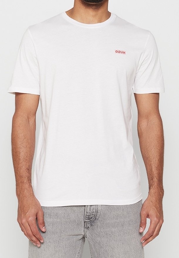 T-shirt en coton blanc à manches courtes, décolleté rond et petit logo rouge sur la poitrine gauche. Porté avec un jean gris clair.
