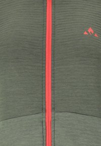 Grüne Stoffjacke mit einem strukturierten Muster, ausgestattet mit einem zentralen roten Reißverschluss und einem kleinen Logo in der oberen rechten Ecke.