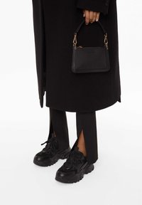 Sac à main en cuir noir avec des accessoires en or, tenu dans la main. Portant des baskets noires avec une surface texturée et une semelle épaisse, associées à un pantalon noir à fente.