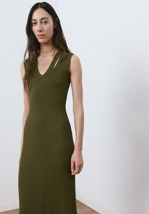 Femme aux longs cheveux bruns portant une robe sans manches en maille côtelée vert olive avec un col en V et une découpe asymétrique, sur un fond blanc uni.