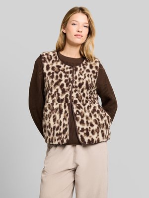 Jeune femme portant un pull marron, un pantalon beige et un gilet sans manches en peluche avec un motif léopard marron et crème.