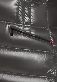 Zwart glanzende pufferjack van stof met een doorgestikt ontwerp. Beschikt over een ritszak met zwarte hardware en een klein rood logo-label.