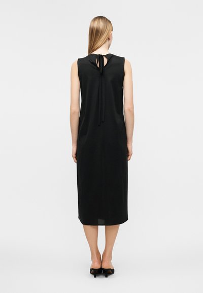 Γυναίκα με μακριά ξανθά μαλλιά, φορεμένη με μαύρο midi φόρεμα χωρίς μανίκια με δέσιμο στην πλάτη, στέκεται με πλάτη, σε μαύρα τακούνια.