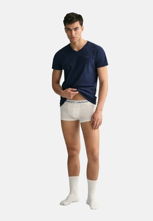 Young man wearing navy blue T-shirt, white boxer briefs, and white socks, lifting shirt slightly to reveal waistband.