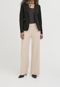 Veste noire cintrée, associée à un pantalon beige à jambes larges. La tenue comprend un haut foncé et une écharpe à motifs nouée au cou.