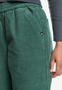 Green corduroy pants with a ribbed texture, elastic waistband, side pocket, and a small black logo patch on the left leg.