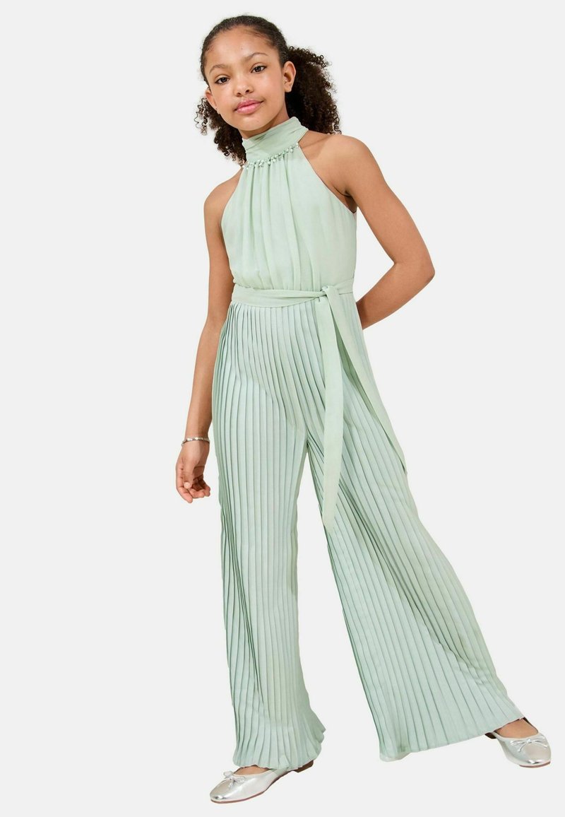 Jumpsuit plissé vert clair avec un col halter et un détail de taille nouée, présentant une texture lisse et une silhouette fluide. Ballerines argentées.