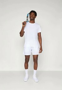 Balta sportinė marškinėliai ir šortai, pagaminti iš kvėpuojančio audinio. Modelis laiko teniso raketę, vilkintis baltus sportinius batelius ir kojines.