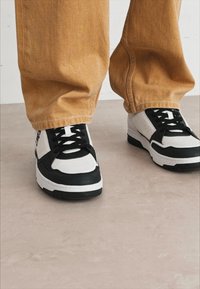 Sneaker bianco e nero con finitura in pelle testurizzata, dotato di punta arrotondata, suola spessa e lacci neri, indossato con pantaloni beige.