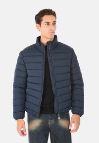 Giubbotto puffer di colore navy con cuciture orizzontali, colletto alto e due tasche con zip. Indossato sopra un maglione nero e abbinato a jeans sbiaditi.