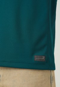 Camisa polo de tela texturizada en color verde azulado con una etiqueta negra de texto blanco, dobladillo cosido, usada sobre pantalones beige claro.