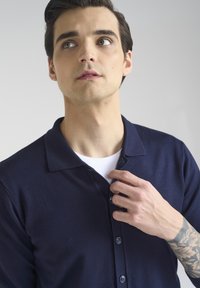 Granatowa koszulka polo z kołnierzykiem, z zapięciem na guziki i plecioną teksturą, noszona na białej koszulce. Gładkie wykończenie materiału.