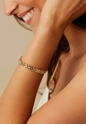 Goldene Kettenarmband an einem gebräunten Handgelenk, mit miteinander verbundenen Gliedern und einer glatten Oberfläche, die die reflektierende metallische Textur betont.