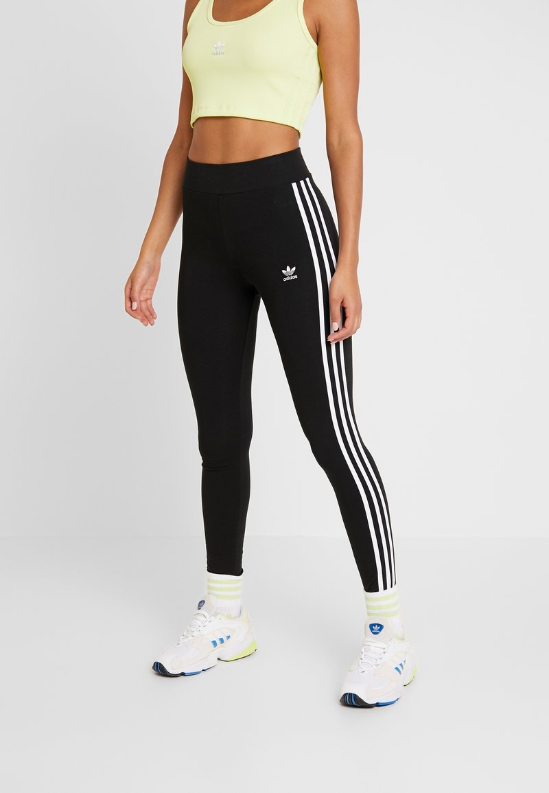 Mustat legginsit, joissa on kolme valkoista raitaa sivuilla ja Adidas-logo. Yhdistetty vaaleankeltaiseen lyhythihaiseen yläosaan ja valkoisiin lenkkareihin.