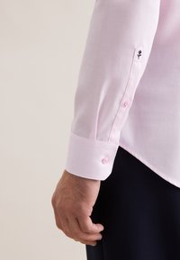 Camicia rosa chiaro a maniche lunghe con tessuto strutturato, polsini con bottoni rosa e un piccolo dettaglio floreale ricamato sulla manica.