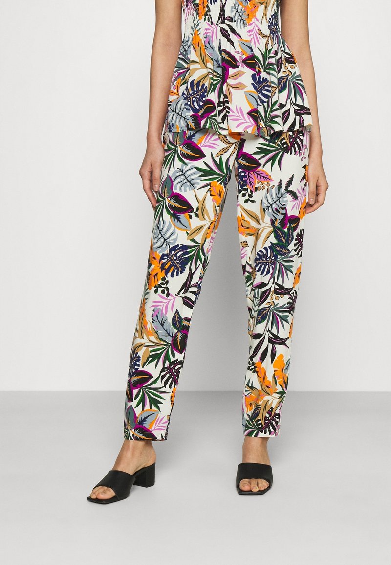 Femme portant un pantalon à imprimé floral coloré et un haut assorti, associée à des sandales à talon bloc noir à bout ouvert, se tenant devant un fond uni.