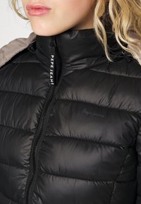 Černá vycpaná bunda s vysokým límcem, s plným zipem a lesklou texturou. Obsahuje jemné značkování na přední straně a na rameni.