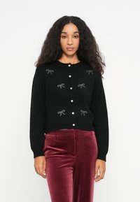 Femeie cu păr creț poartă un cardigan negru cu fundi�țe din strasuri și butoni albi, combinat cu pantaloni din catifea bordeaux, stând pe un fundal alb.