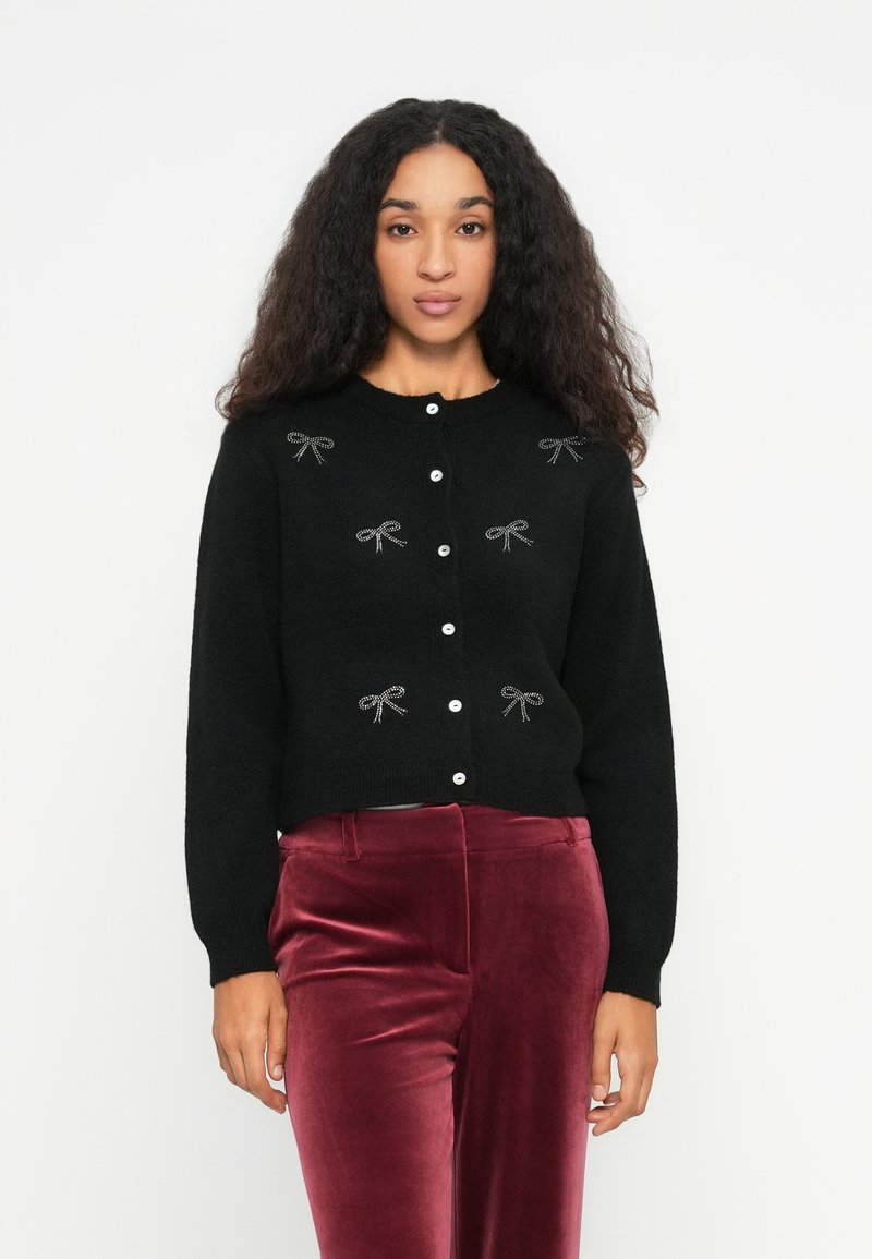 Femeie cu păr creț poartă un cardigan negru cu fundițe din strasuri și butoni albi, combinat cu pantaloni din catifea bordeaux, stând pe un fundal alb.