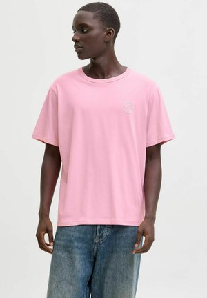 Persona joven con camiseta rosa de manga corta con pequeño logo blanco y vaqueros azules, mirando hacia un lado contra un fondo liso.