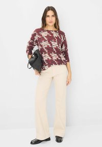 Maglione a motivi rossi e crema con un design geometrico, abbinato a pantaloni beige a zampa d'elefante e scarpe nere, completato da una borsa nera.