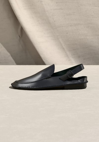 Chaussure plate slingback en cuir noir avec détails tissés et bride à boucle réglable, photographiée sur une surface beige avec un fond en tissu.