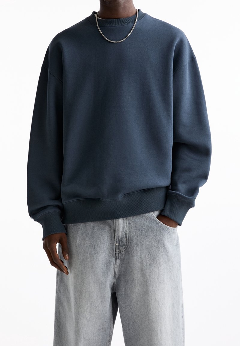 Personne portant un sweat-shirt à col rond bleu foncé, un jean gris clair, un collier chaîne en argent, avec une main dans la poche sur un fond blanc.