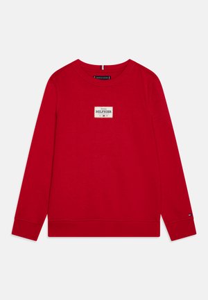 Piros pulóver hosszú ujjal, kerek nyakkivágással és bordázott mandzsettával. Az elején egy téglalap alakú logó címke található a márkanevével.