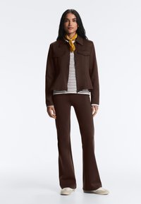 Woman standing wearing brown jacket and pants, striped shirt, yellow neck scarf, and white sneakers on white background.