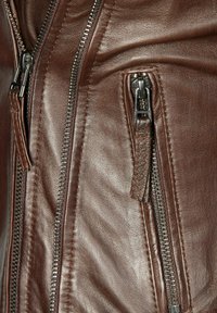 Braune Lederjacke mit mehreren silbernen Reißverschlüssen, strukturierter Oberfläche und genähten Details an den Nähten für zusätzliche Haltbarkeit.