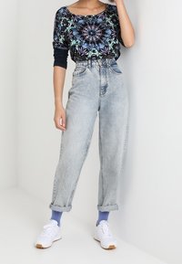 Una persona che indossa una maglietta a maniche lunghe con un motivo, jeans a vita alta di un blu chiaro arrotolati alle caviglie, calze blu e sneakers bianche.