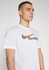 Camiseta de algodón blanca con un logo de Nike naranja y el texto "Running" en negro, con mangas cortas y cuello redondo.