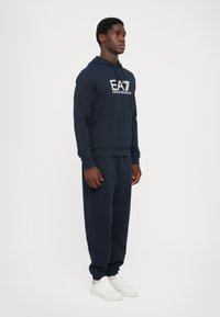 Felpa in cotone blu navy con tasca a marsupio frontale e logo, abbinata a pantaloni jogger coordinati e scarpe da ginnastica bianche, con polsini elasticizzati.